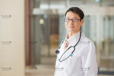 病院内に立つ聴診器を首にかけ眼鏡をかけた男性医師の画像