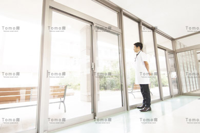 病院の廊下に立ち外を眺める男性医師の画像