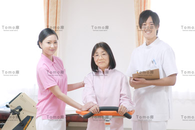 リハビリ室に立つ笑顔の男性看護師と患者と女性理学療法士の画像