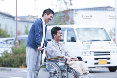 駐車場を移動する車椅子の男性患者と男性医療スタッフの画像