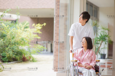 病院の中庭を眺める車椅子の女性患者と男性看護師の画像