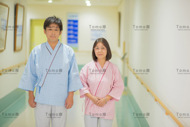 病院の廊下に立つ検査着の患者2名・健康診断/検査