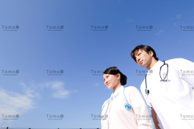 青空を背景に立つ男性医師と女性看護師（ナース）の画像