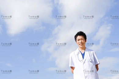 青空を背景に笑顔で立つ若い男性医師の画像