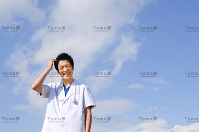 青空を背景に笑顔で立つ若い男性医師の画像